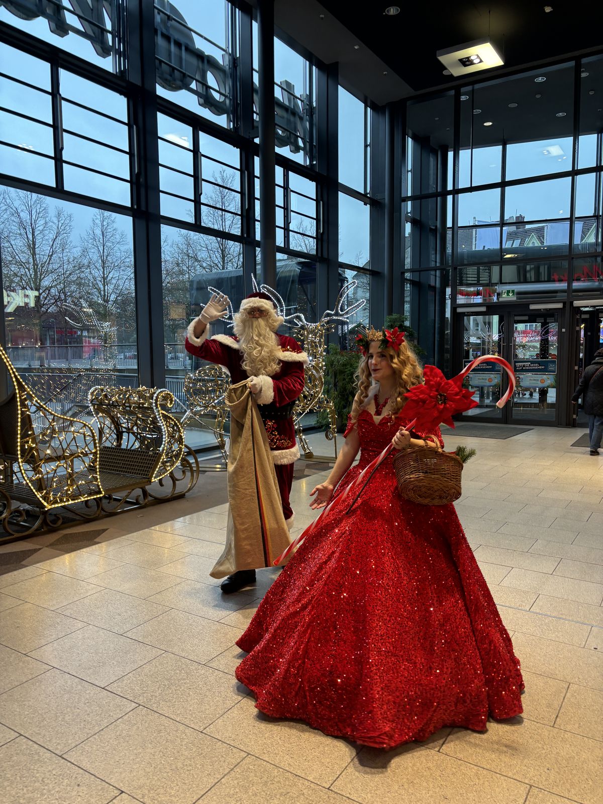 Weihnachtsmann und Weihnachtsengel laufen an goldenen Schlitten vorbei durch ein Einkaufszentrum