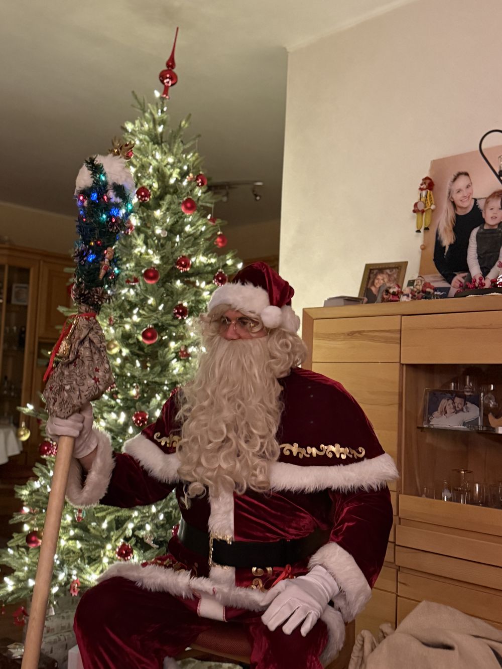 Der Weihnachtsmann sitzt mit Stab im Wohnzimmer neben dem Weihnachtsbaum